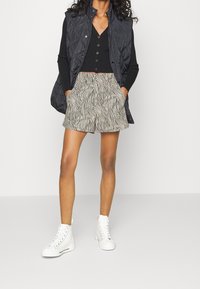 Kvinna i svart långärmad topp, svart vadderad väst, shorts med zebramönster och vita högklackade sneakers som står med händerna i fickorna.