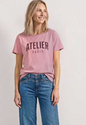 Femme portant un T-shirt rose avec le texte "ATELIER DE PARIS" et un jean bleu, souriante et regardant sur le côté sur un fond uni.