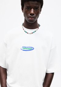 White oversized t-shirt featuring "CREATIVE MINDSET" graphic in blue and green, paired with a multicolored beaded necklace.
