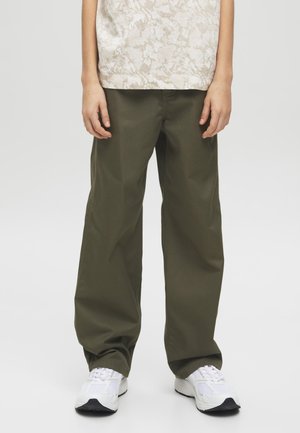 Persoană care poartă pantaloni largi verde-olive, adidași albi și o cămașă ușoară cu model, stând în fața unui fundal simplu.