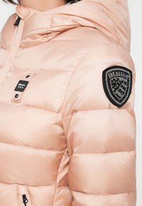 Lichtroze pufferjack met capuchon, voorzien van een zwart logo-patch op de linkermouw, een gladde textuur en een drukknoopsluiting aan de voorkant.