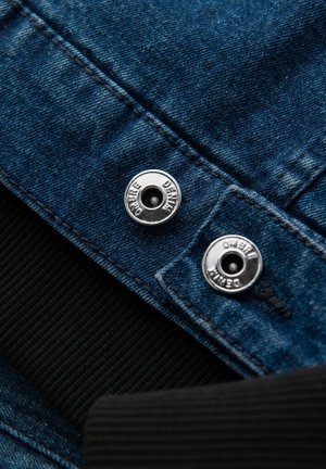 Jeansjacka med blått tyg, silverfärgade tryckknappar märkta med "ombre denim", och en svart ribbad krage.
