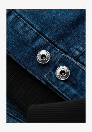 Chaqueta de denim con tela azul, botones de presión plateados marcados con "denim ombre" y un puño de cuello acanalado negro.