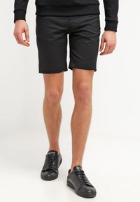 Schwarze, maßgeschneiderte Shorts aus glattem Stoff, mit schmaler Passform und umgeschlagenen Säumen. Kombiniert mit schwarzen Sneakers, die ein minimalistisches Design aufweisen.