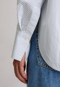 Fehér ing kék csíkokkal, gombos mandzsettával és oldalsó nyílással, farmerrel párosítva. Látható a kar és az anyag textúrája.