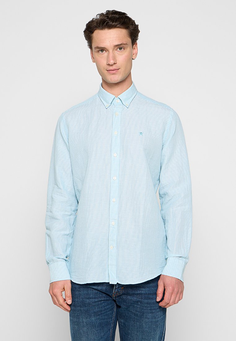 Junger Mann in einem hellblauen Hemd mit Knöpfen und kleinem Logo auf der Brust, sowie dunkelblauen Jeans, der vor einem schlichten Hintergrund steht.