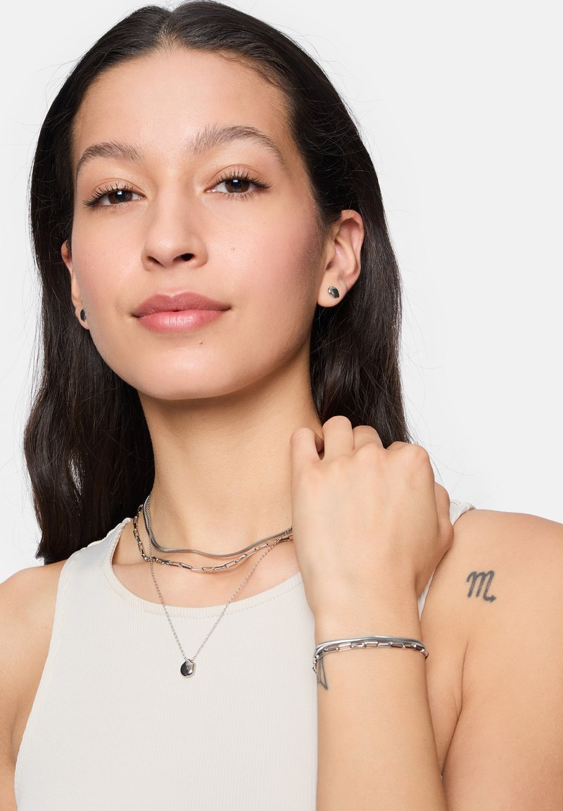 Zilveren sieraden met meerdere gelaagde kettingen en een armband, geplaatst op een lichte achtergrond, met een close-up van huiddetails en tatoeages.