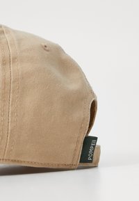 Beige Baseballcap aus strukturiertem Stoff, mit einer gesteppten Naht und einem grünen Etikett an der Seite, auf dem "POMPEII" steht.