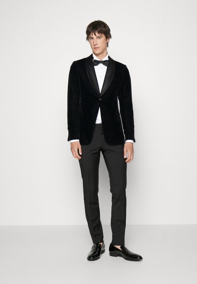 Veste de smoking en velours noir avec revers en satin, associée à une chemise blanche et un pantalon noir. Des chaussures de ville noires brillantes complètent le look.
