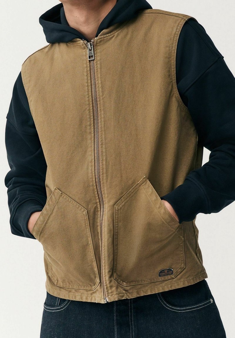 Chaleco de lona color canela con cierre frontal de cremallera, grandes bolsillos angulados y sudadera negra con capucha debajo. Detalles visibles de costuras y herrajes.