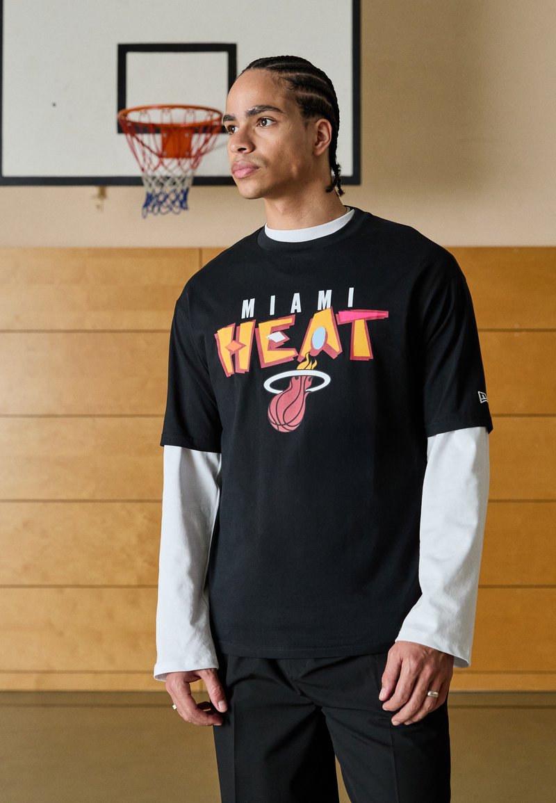 Camiseta negra con un atrevido gráfico "MIAMI HEAT" en letras coloridas y el logotipo, superpuesta a una camisa de mangas largas blanca, combinada con pantalones negros.