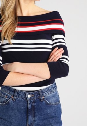 Femme portant un pull bleu marine à épaules dénudées avec des rayures horizontales blanches et rouges, bras croisés, associé à un jean bleu taille haute.