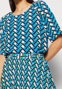 Blå mønstrede top med et vævet design i nuancer af blå, grøn og sort; letvægtsstof, rund halsudskæring og korte ærmer.