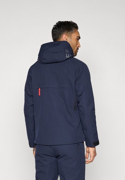 Tamsiai mėlyna neperšlampama striukė su gaubtu, turi raudoną akcentinį kilpą, du šoninius užtrauktukus ir matomą logotipą gale.