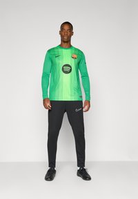 Maglia da portiere di un brillante colore verde con strisce verticali, caratterizzata da un logo centrale di Spotify in nero. Indossata con pantaloni sportivi neri Nike.