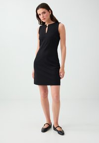 Woman wearing a sleeveless black dress with a keyhole neckline and gold button, paired with black Mary Jane flats, standing on a white floor.
