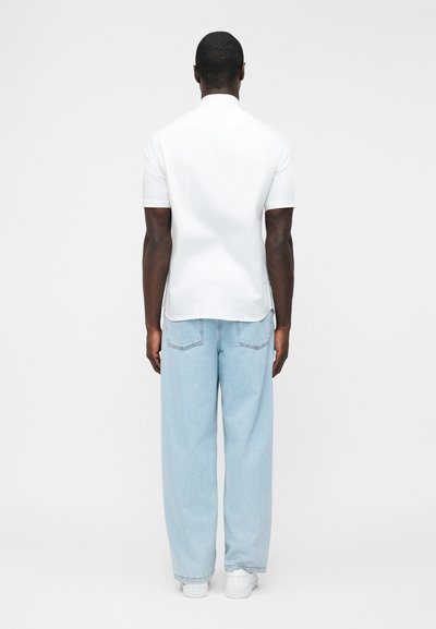 Άνδρας που στέκεται με την πλάτη, φοράει λευκό πουκάμισο με κοντά μανίκια, ανοιχτό μπλε τζιν με φαρδιά μπατζάκια και λευκά αθλητικά παπούτσια σε απλό φόντο.