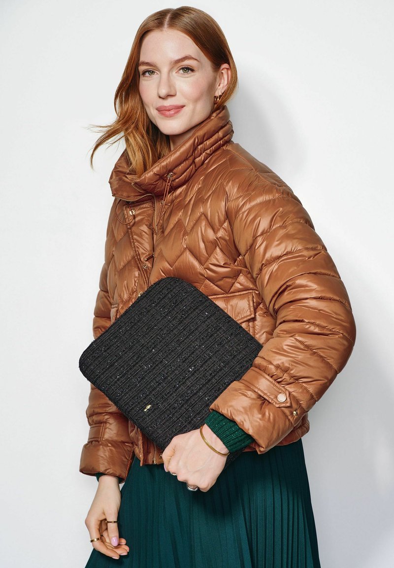 Brązowa puchowa kurtka z wysokim kołnierzem, czarna teksturowana kopertówka, plisowana zielona spódnica oraz metaliczne akcenty na kurtce i biżuterii.