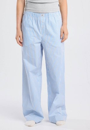Persoon in loszittende blauw-witte verticaal gestreepte pyjamabroek met een grijs shirt en witte sokken, staand tegen een effen achtergrond.