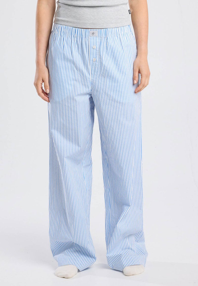 Persona vistiendo pantalones de pijama holgados de rayas verticales azules y blancas, con una parte superior gris y calcetines blancos, de pie contra un fondo simple.