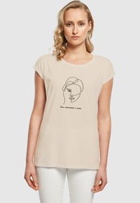 Beige bomulds t-shirt med korte ærmer og rund halsudskæring. Har en minimalistisk sort linjetegning af et ansigt og trykt tekst.