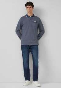 Granatowa koszulka polo z długim rękawem, białym kołnierzykiem i detalem na kieszeni, zestawiona z niebieskimi jeansami i białymi tenisówkami, stojąca na neutralnym tle.