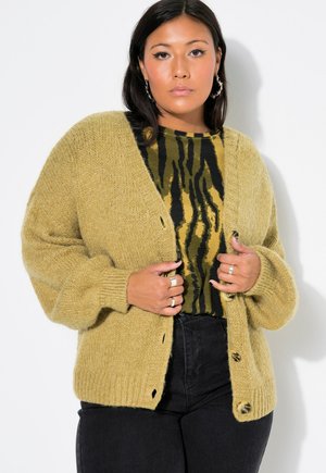 Cardigan a maglia in giallo tenue, con una vestibilità ampia, polsini a coste e grandi bottoni, indossato sopra una blusa a fantasia verde e nera.