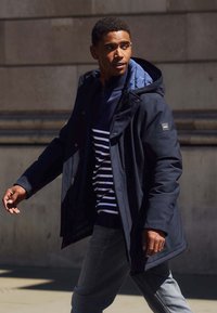 Veste extérieure marine avec capuche et fermeture éclair à l'avant, superposée à un haut rayé bleu marine et blanc, assortie à un jean gris. Style urbain décontracté.