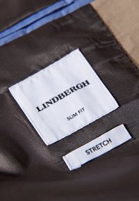 Schwarzes Innenfutter mit weißen und schwarzen Etiketten mit der Aufschrift "LINDGRGH", "SLIM FIT" und "STRETCH" vor einem strukturierten Stoffhintergrund.