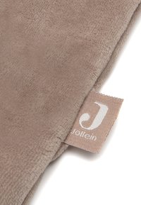 Jollein zacht beige fleece deken met een gladde textuur, voorzien van een gestikt label met een logo in het wit. Rechthoekige vorm, lichtgewicht ontwerp.