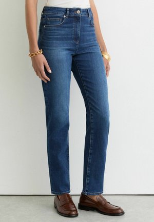 Mujer que lleva vaqueros azules de pierna recta, mocasines de cuero marrón, una camiseta blanca, una pulsera de cadena de oro y una pulsera rígida dorada frente a una pared lisa.