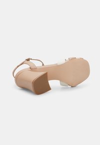 Sandale à talons hauts beige avec des brides accentuées blanches. Elle présente un large talon carré et une semelle en caoutchouc texturée pour une meilleure adhérence. Fermeture à boucle à la cheville.