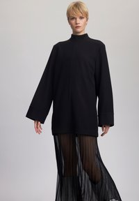Fekete, oversize méretű pulóver magas nyakkal, hosszú ujjakkal és minimalista dizájnnal, egy áttetsző fekete, pliszírozott szoknya fölé rétegezve.