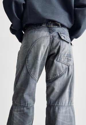 Vue de dos d'une personne portant un jean gris délavé avec des coutures courbes et une poche à rabat boutonnée sur la hanche droite, associée à un sweat-shirt bleu marine.