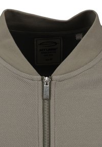 Felpa grigia con zip, caratterizzata da una texture a coste, colletto alto e una zip in metallo con un accento verde. L'etichetta indica la taglia M.