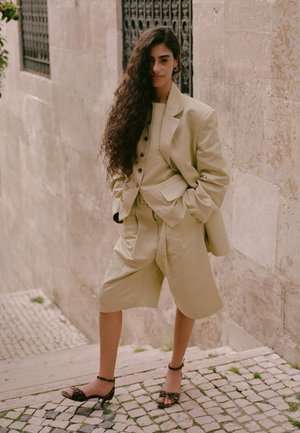Jeune femme aux longs cheveux bouclés portant un costume beige oversize avec un short et des talons imprimé léopard, debout sur des marches en pierre à l'extérieur.