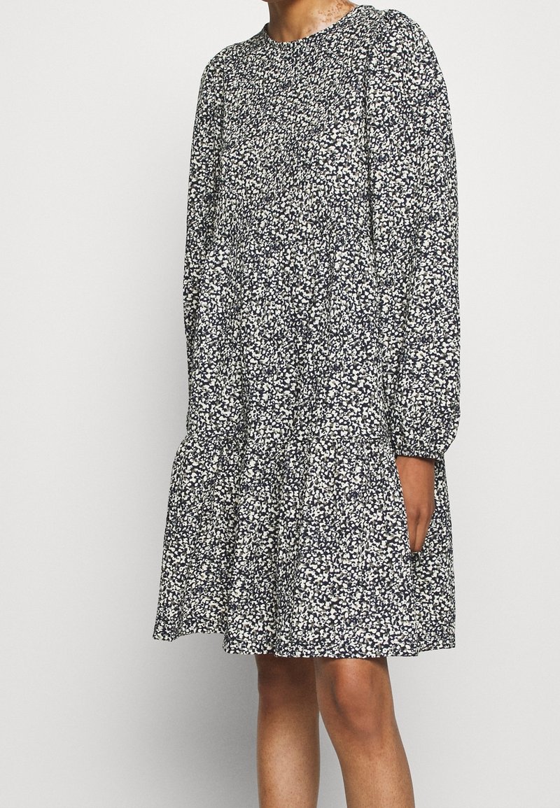 Robe à motif floral en bleu marine et blanc, avec un col rond, des manches longues et une jupe évasée à volant. Tissu léger.