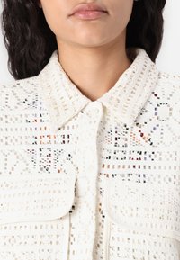 Biała bluzka z szydełkowanej tkaniny z wyraźnym kołnierzykiem, zapięciem na guziki i dwiema przednimi kieszeniami; cechuje ją geometrzyczny wzór z kolorowymi nićmi.