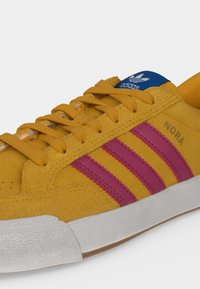 Κίτρινο καστόρινο αθλητικό παπούτσι Adidas με ροζ ρίγες, λευκή σόλα, μπλε ετικέτα με λογότυπο και "NORA" τυπωμένο στο πλάι.
