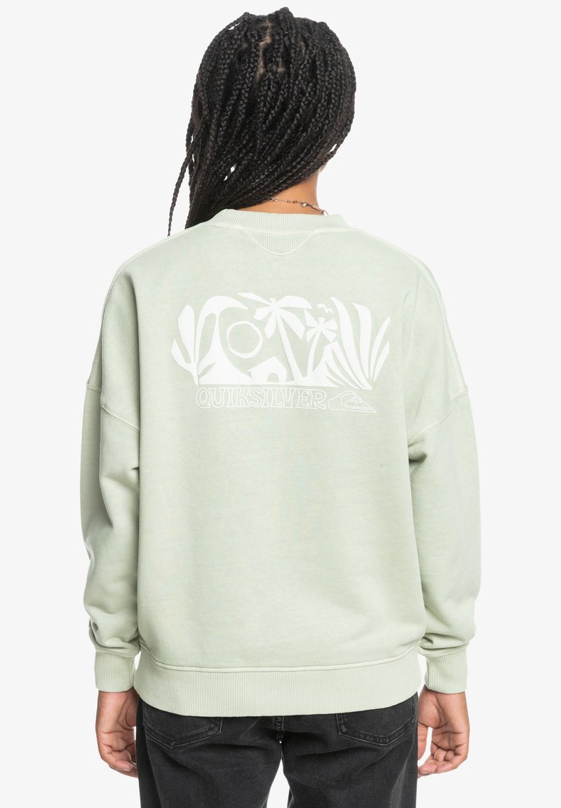 Lys grønn crewneck sweatshirt laget av myk stoff. Har et stort hvitt grafisk trykk på baksiden, inkludert tropiske motiver og merkenavn.