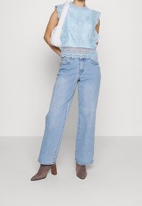 Ljusblå spetsblus med vågformade kanter, kombinerad med lösa, raka ljusa denimjeans. Modellen håller en fluffig vit handväska. Bruna klackboots.