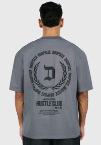 Grå oversized t-shirt lavet af bomuld med et stort trykt design på ryggen, der har tekst og et cirkulært grafisk motiv.