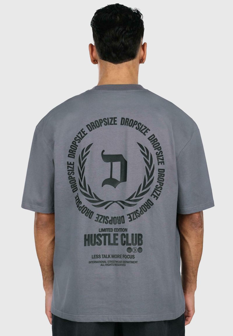 Grå oversized t-shirt lavet af bomuld med et stort trykt design på ryggen, der har tekst og et cirkulært grafisk motiv.
