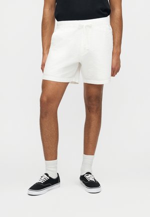 Άνδρας που φοράει λευκά, υφασμάτινα σορτς με κορδόνια, συνδυασμένα με λευκές κάλτσες και μαύρα αθλητικά παπούτσια χαμηλού ύψους, μπροστά σε απλό φόντο.