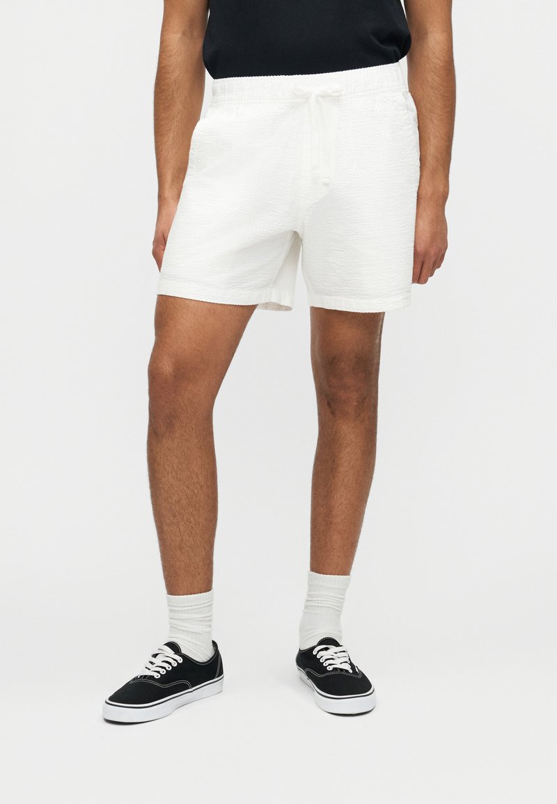 Άνδρας που φοράει λευκά, υφασμάτινα σορτς με κορδόνια, συνδυασμένα με λευκές κάλτσες και μαύρα αθλητικά παπούτσια χαμηλού ύψους, μπροστά σε απλό φόντο.