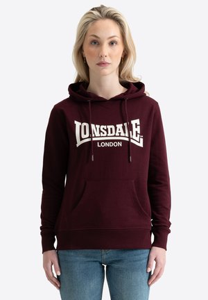 Blondide juustega naine, kes kannab burgundia värvi Lonsdale Londoni kapuutsiga pusa ja siniseid teksaseid, seisab hele taustaga eespool.