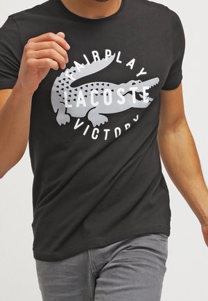 Sort bomuld T-shirt med et gråt krokodillemotiv og teksten "FAIRPLAY LACOSTE VICTORY" i fede hvide bogstaver. Korte ærmer, rund hals.