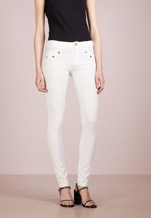 Kvinde iført hvide slim-fit bukser med dekorative knapper og sorte højhælede sandaler med ankelremme, stående mod en neutral baggrund.
