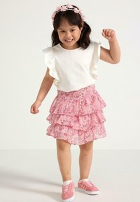Top bianco a maniche corte con bordi arricciati, abbinato a una gonna rosa a strati con motivi floreali, e sneakers rosa con suola bianca.