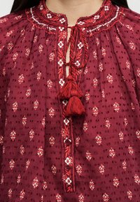 Gros plan sur un chemisier rouge à petits motifs floraux, col brodé, patte de boutonnage et deux pompons rouges noués au niveau du cou.